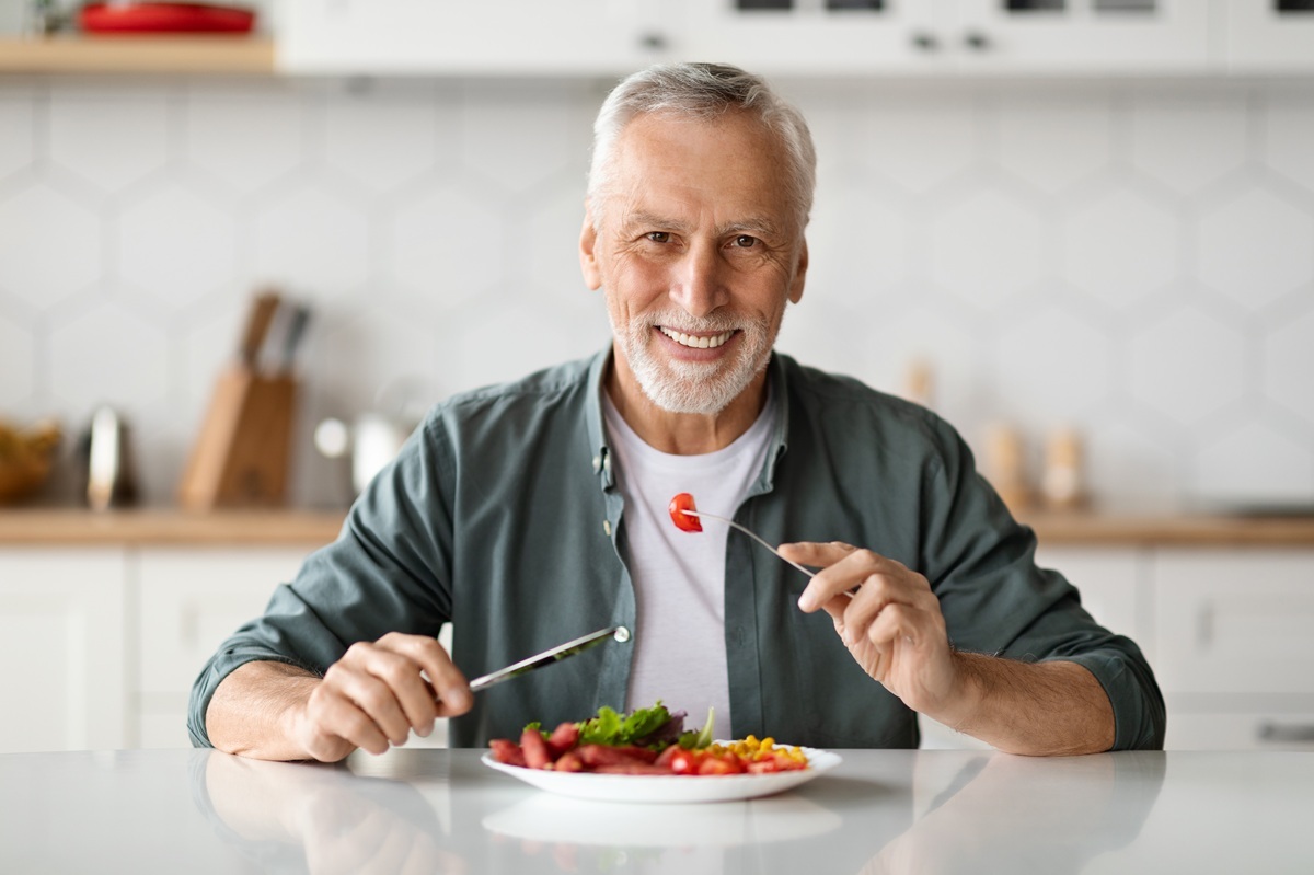 Smiling mature man enjoying healthy salad in kitchen, representing post-implant dietary recovery.